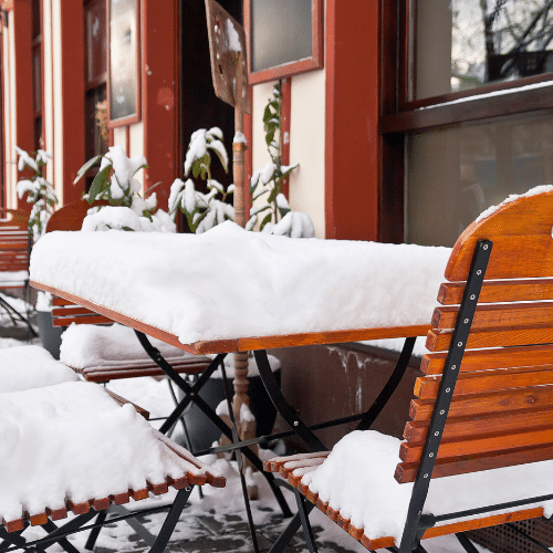 Buiten terras met houten tafels en stoelen bedekt met een laag sneeuw in een winterse omgeving.