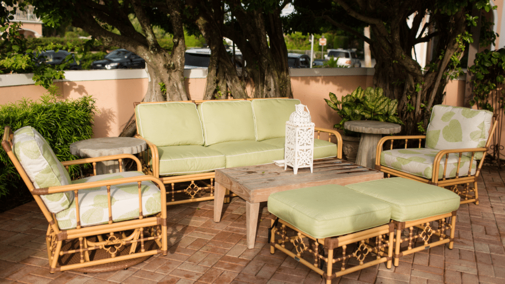 Luxe bamboe tuinset met groene kussens en houten salontafel, geplaatst op een betegeld terras met een achtergrond van grote bomen en een sfeervolle tuinmuur.