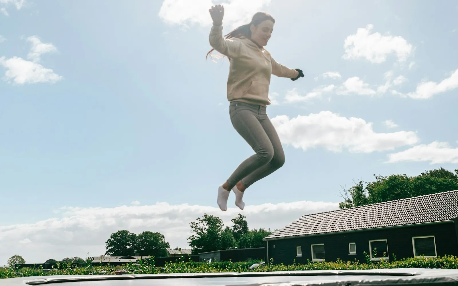 Woman jumping high above a trampoline in a garden, with a house and trees in the background